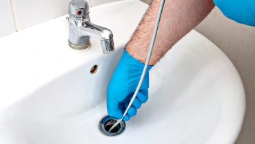 A plumber using a drain snake to unclog a deep clogged pipe beneath a sink drain.