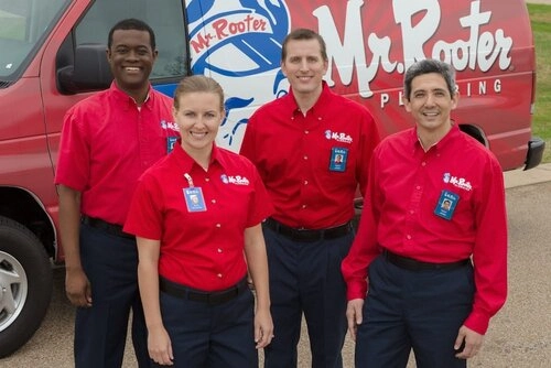 Four smiling plumbers wearing Mr. Rooter Plumbing uniforms and standing in front of a van branded with the Mr. Rooter Plumbing logo.