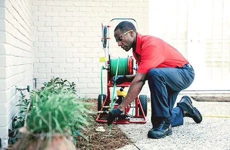 A plumber from Mr. Rooter Plumbing kneeling near a drain opening in a garden while lowering a tube into the opening from a nearby coil for the purposes of hydro jetting.