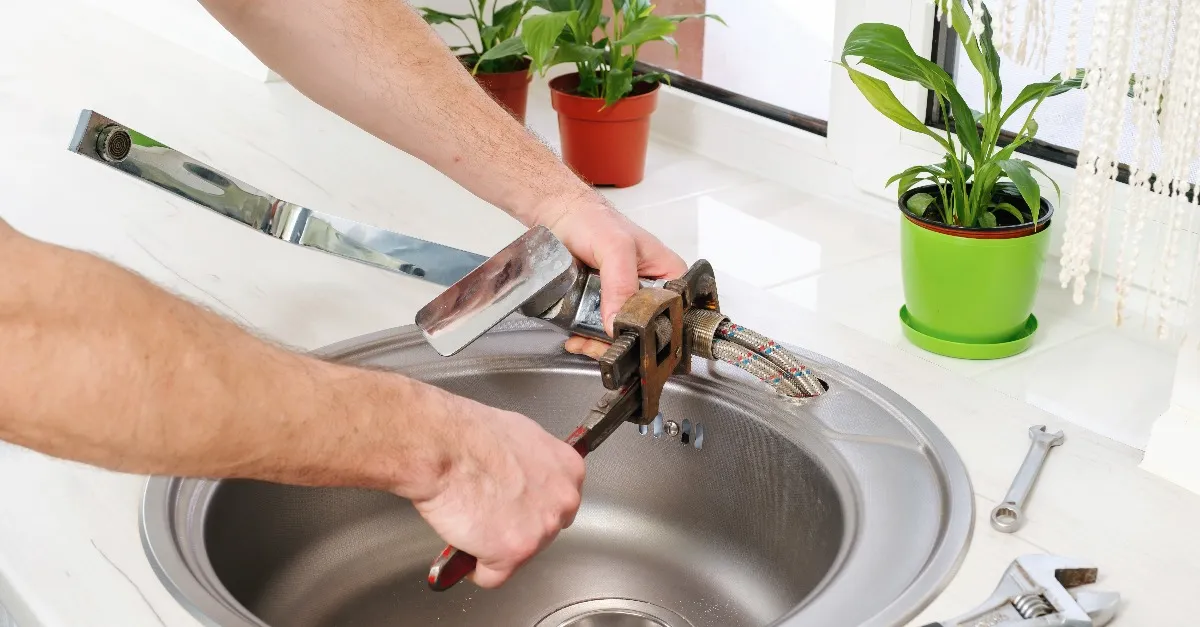 A plumber using a wrench to fix a faucet that has been partially detached from a sink.