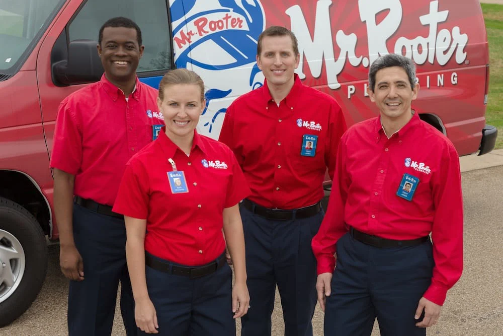 A group of smiling plumbers dressed in Mr. Rooter Plumbing uniforms and standing in front of a branded work van.