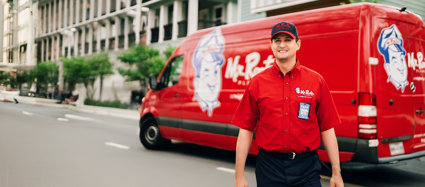 Mr. Rooter technician carrying mat and utility bag in front of branded red work van.