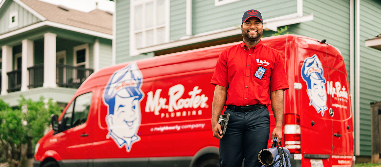 Mr. Rooter technician carrying mat and utility bag in front of branded red work van.