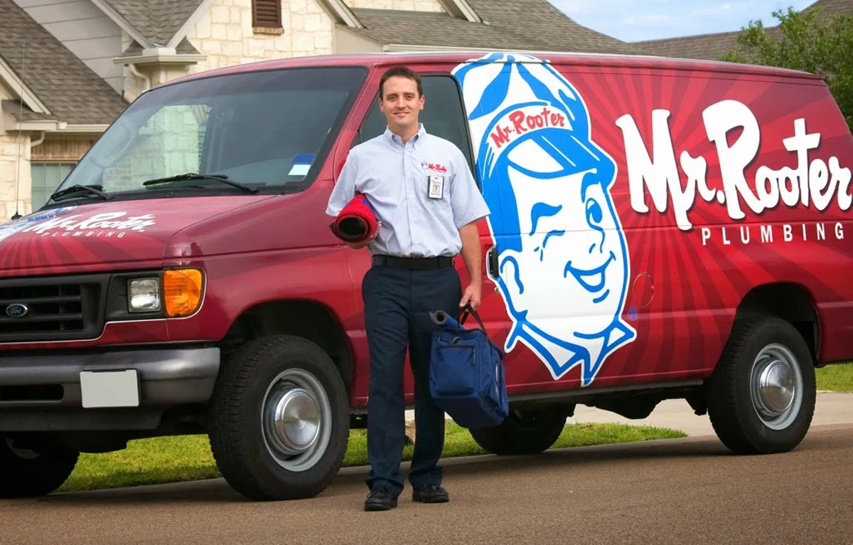 A plumber wearing a Mr. Rooter Plumbing uniform and carrying tools from a van branded with the Mr. Rooter Plumbing logo.