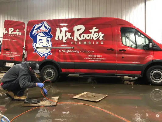 A plumber in Sherwood park lowering a sewer scope into a floor drain with a large, branded Mr. Rooter Plumbing van visible in the background.